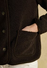 Hnedá flísová bunda s textúrovaným povrchom, s gombíkovým zapínaním, náprsním vreckom a kontrastnými kordovými okrajmi.