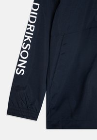 Tumši zila jaka ar elastīgām aprocēm, uz piedurknes redzams izteiksmīgs balts "DIDRIKSONS" logotips, gluda auduma tekstūra un šuvju detaļas.