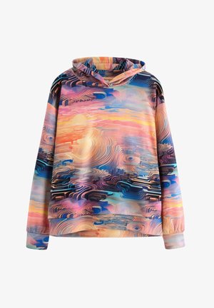 Tienerjongen lopend, gekleed in een hoodie met een meerkleurig abstract patroon, crème broek en witte sneakers, tegen een effen lichte achtergrond.