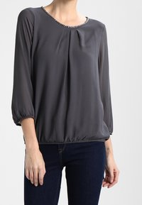 Graue Bluse mit Dreiviertelärmeln, gefältelter Vorderseite und einem abgerundeten Ausschnitt, verziert mit einem dekorativen Besatz. Weiche, leichte Stoffqualität.