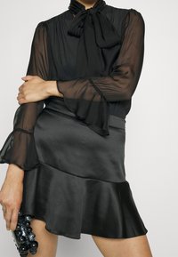 Black sheer blouse with long ruffled sleeves and a tie at the neck, paired with a black satin flared mini skirt, held with one hand.