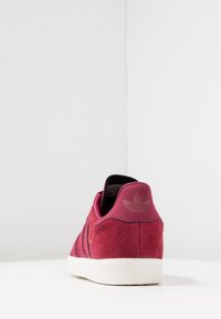 Röd mockaskor med svarta ränder, texturerad baksida och liten logotyp. Den kontrasterande vita gummisulans ger hållbarhet och grepp.