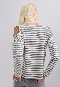 Mujer con cabello rubio largo que lleva una blusa a rayas blanca y negra con los hombros al aire y pantalones grises, con las manos en los bolsillos traseros.
