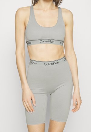 Kobieta nosząca jasno szary prążkowany sportowy biustonosz Calvin Klein oraz dopasowane, wysoko taliowane szorty rowerowe na neutralnym tle.