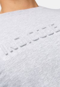 Camiseta de algodón gris con un diseño elevado, texturizado y en relieve que presenta la palabra "INCLUSIVO" en el pecho. Tejido de punto suave y liso.