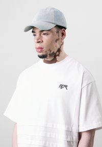 Light blue denim cap, white oversized t-shirt with layered texture and black "ADR" logo. Individual has a distinct skin pattern.