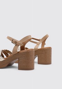 Sandalias de tacón bloque beige con una correa de textura trenzada, parte superior metálica y hebilla ajustable en el tobillo. Diseño elegante con materiales suaves.