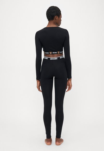Naine seisab paljajalu seljaga pööratud, kandes musta pikakätist lühikest toppi ja leggings'eid, mille vöö ja äär on Moschino kaubamärgiga elastsete ribadega.