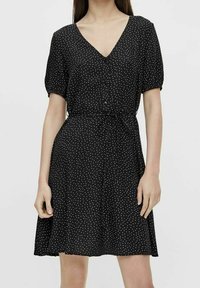 Czarna sukienka w białe kropki, krótkie bufiaste rękawy, dekolt w kształcie litery V, guziki z przodu i zwężona talia prowadząca do rozkloszowanej spódnicy.