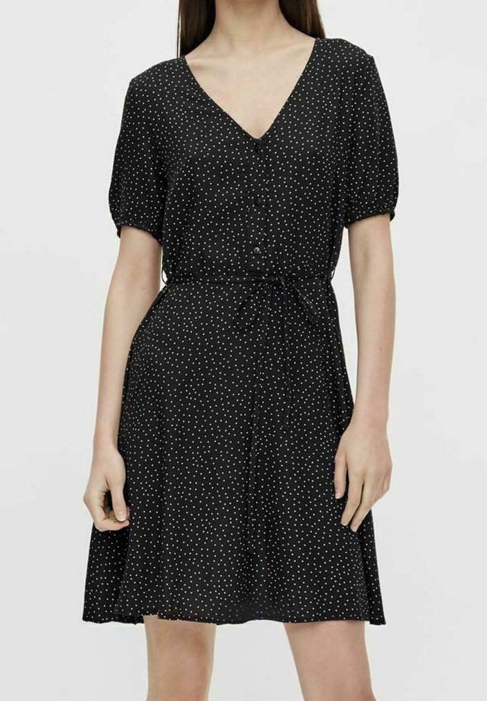 Czarna sukienka w białe kropki, krótkie bufiaste rękawy, dekolt w kształcie litery V, guziki z przodu i zwężona talia prowadząca do rozkloszowanej spódnicy.