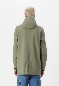 Khaki nepromokavá bunda s vysokým límcem a zadním větracím otvorem, s hladkou texturou a dlouhými rukávy, v kombinaci s modrými cargo kalhotami.