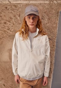 Lys beige zip-up jakke med høj krave, grå T-shirt under, og matchende grå kasket. Casual pasform med elastiske manchetter og kant.