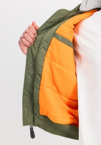 Giacca verde trapuntata con fodera arancione, che presenta una tasca con zip e polsini a costine. Una mano si afferra all'apertura, rivelando la texture interna.