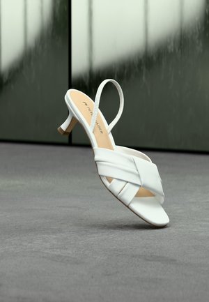 Λευκό σανδάλι με τακούνι, με λουριά που διασταυρώνονται και ιμάντα πίσω από το πόδι, τοποθετημένο όρθιο πάνω σε γκρι επιφάνεια, με σκούρο θολωμένο φόντο.