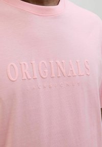 Gaiši rozā T-krekls ar apaļu kakla izgriezumu, izgatavots no kokvilnas, ar reljefiem uzrakstiem "ORIGINALS" un "JACK & JONES" nedaudz tumšākā nokrāsā.