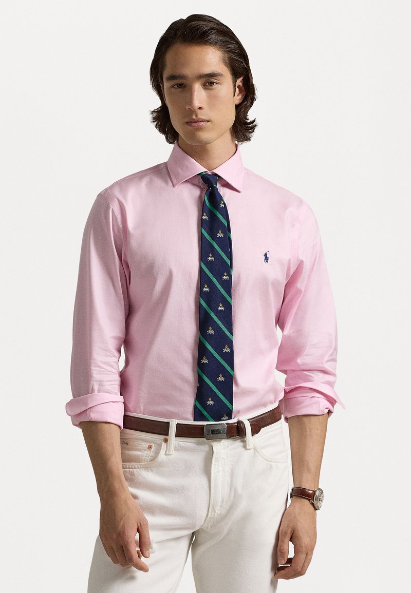 Camisa rosa de manga longa com as mangas dobradas, apresentando uma gravata azul marinha com riscas verdes e pequenos emblemas, combinada com calças brancas e um cinto castanho.