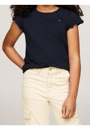 Barn iført en marineblå t-shirt med korte ærmer og flæsekant samt cremefarvede cargobukser med lommer, stående med den ene hånd på hoften.