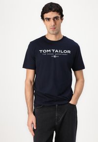 Tamsiai mėlynas medvilninis marškinėliai su apvaliu kaklu, su balta spauda: "TOM TAILOR THE CASUAL DEPARTMENT 1962." Dėvimi su juodais džinsais.