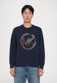 Sweatshirt azul-marinho com um gráfico redondo apresentando o logótipo "MK" em cores metálicas. Gola redonda e punhos canelados, feito de um material têxtil suave.
