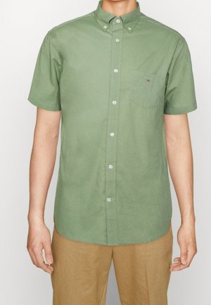 Homme portant une chemise verte à manches courtes boutonnée avec une poche poitrine et un pantalon beige, debout devant un fond uni.