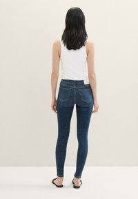 Tijesne traperice u tamnoplavoj denim boji, s srednje visokom strukom, dva stražnja džepa i uskim krojem, uparene s bijelim topom bez rukava.