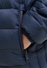 Marinblå pufferjacka med quiltat mönster, med en kontrasterande röd dragkedja och en svart dragkedjehandtag, där en hand sträcker sig in i en ficka.
