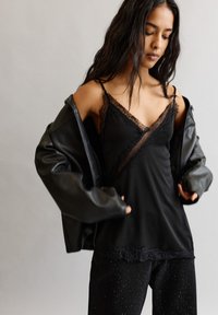 Nő fekete bőrdzsekijét igazítja a fekete csipkével díszített topja és a diszkrét fényes fekete nadrágja felett, egyszínű háttér előtt.