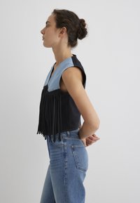 Schwarzes Crop-Top mit Denim-Akzenten, das Fransen an der Vorderseite und Druckknöpfe aufweist, kombiniert mit hochgeschnittenen blauen Jeans.
