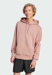 Felpa rosa chiaro con tasca frontale e cappuccio con cordino, con maniche lunghe e un logo sul braccio sinistro. Abbinata a pantaloni corti neri.