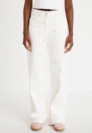 Hvide bukser med vide ben og små pink og gule tusindfryd printet, båret af en person, der står, kombineret med beige sneakers og en hvid top.