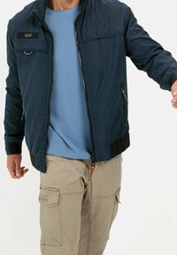 Lekka marynarsko-niebieska kurtka z zamkiem błyskawicznym, bocznymi kieszeniami i ściągaczami na mankietach; noszona na niebieskiej teksturowanej koszuli i beżowych spodniach typu cargo.