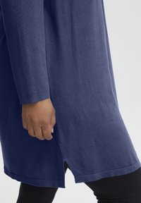 Tunic in maglina blu navy a maniche lunghe con fessure laterali all'orlo, indossata sopra leggings neri. Texture morbida e silhouette rilassata.