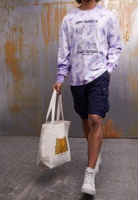 Chemise à manches longues tie-dye violette avec un motif extérieur, associée à un short cargo foncé et des baskets montantes blanches, portant un sac fourre-tout en toile.