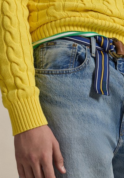 Rumeni pulover s kabelskim vzorcem in rebrastimi manšetami, kombiniran z modrimi raztegnjenimi kavbojkami. Pas z modro-rumeno črtami ima rjavo zaponko.