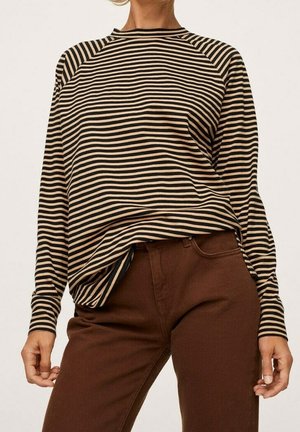 Femme portant un haut à manches longues rayé horizontal noir et beige rentré dans un pantalon marron taille haute, debout devant un fond uni.