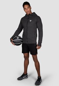 Dark gray zip-up hoodie with a logo, black shorts, and textured sneakers; holding a black and white basketball. Neutral background.