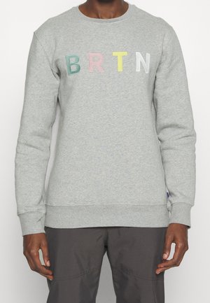 Mężczyzna noszący jasnoszarą bluzę z kolorowymi literami "B R T N" wyhaftowanymi na piersi, połączoną z ciemnymi spodniami.