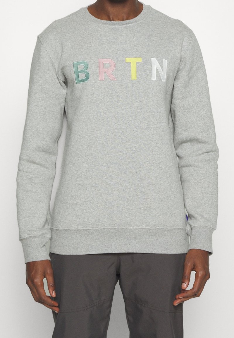 Homme portant un sweat-shirt gris clair avec les lettres colorées "B R T N" brodées sur la poitrine, assorti à un pantalon foncé.