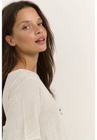 Jasno beżowa lniana bluzka o luźnym kroju, z dekoltem w serek i lekko teksturowanym materiałem. Modelka ma długie, brązowe włosy.