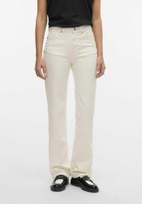Jeans de color crema, de cintura alta y diseño recto, que cuentan con cinco bolsillos y un cierre de botón de metal. Textura suave.
