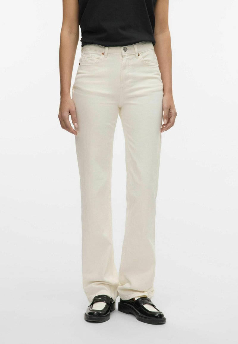 Jeans de color crema, de cintura alta y diseño recto, que cuentan con cinco bolsillos y un cierre de botón de metal. Textura suave.