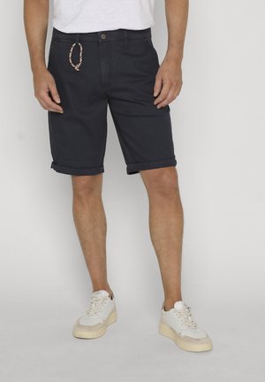 Mężczyzna w czarnych spodenkach z podwiniętymi brzegami, z plecionym brelokiem i białych trampkach, stojący na jednolitym tle.