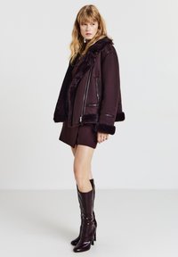 Bordeauxrote Lammfelljacke mit pelzgefüttertem Kragen, durchgehendem Reißverschluss und kontrastierenden Kunstlederakzenten. Das Modell trägt einen passenden Rock und kniehohe Stiefel.