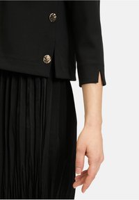 Schwarzer Blazer mit Dreiviertelärmeln, goldenen Knöpfen und geschlitzten Manschetten, kombiniert mit einem plissierten schwarzen Rock, der eine glatte Stoffstruktur präsentiert.