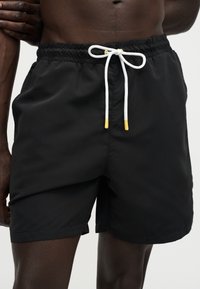 Short de bain noir avec cordon blanc et embouts jaunes porté par une personne à la peau foncée et torse nu, debout avec le bras plié.