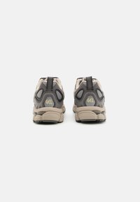 Des chaussures de sport beiges et grises avec une tige texturée, un coussin de talon proéminent, des sangles à l'arrière et un logo « GEL » audacieux sur le talon.