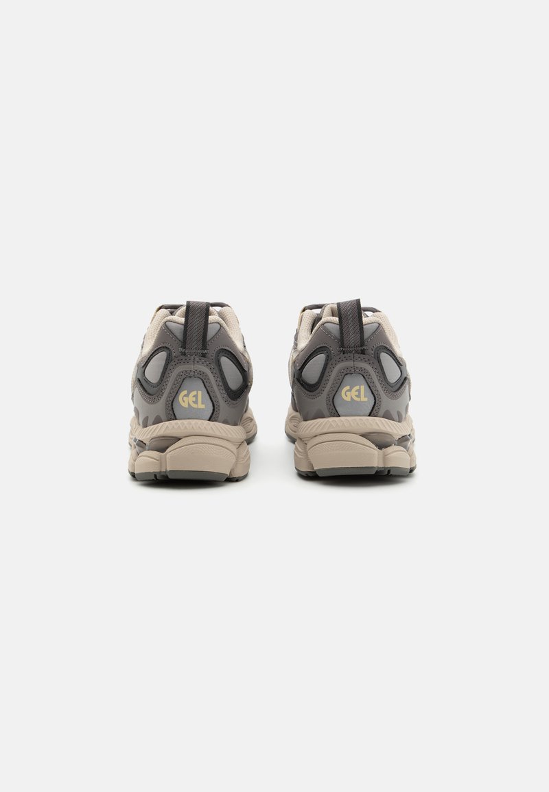 Beige und graue Sportschuhe mit einem strukturierten Obermaterial, ausgeprägter Fersenpolsterung, Rückziehhilfen und einem auffälligen "GEL"-Logo an der Ferse.