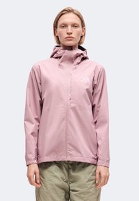 Femme portant une veste à capuche zippée violet clair et un pantalon vert olive, debout, face à l’avant sur un fond uni.