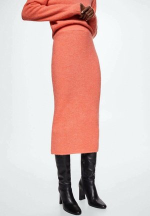 Person iført en tætsiddende koralfarvet strikket nederdel, matchende koralfarvet sweater og sorte knælange støvler med høje hæle, stående med armene over kors.