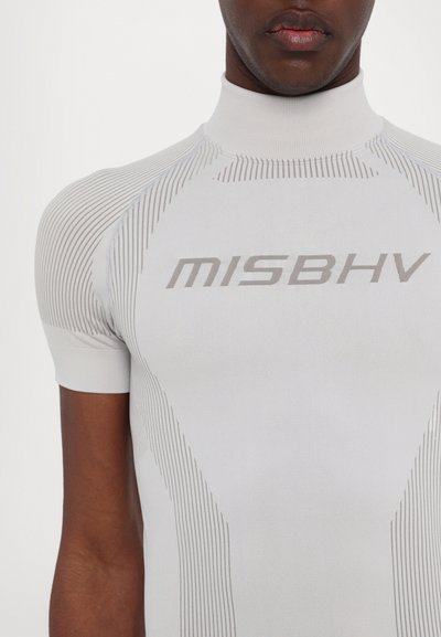 Valge lühikeste varrukatega keha saanud topp, millel on ribakujuline tekstuur ja hallid liniarjuhtmed; esikillegi brändi logo "MISBHV" trükitud ees. Kõrge kaelusega disain.